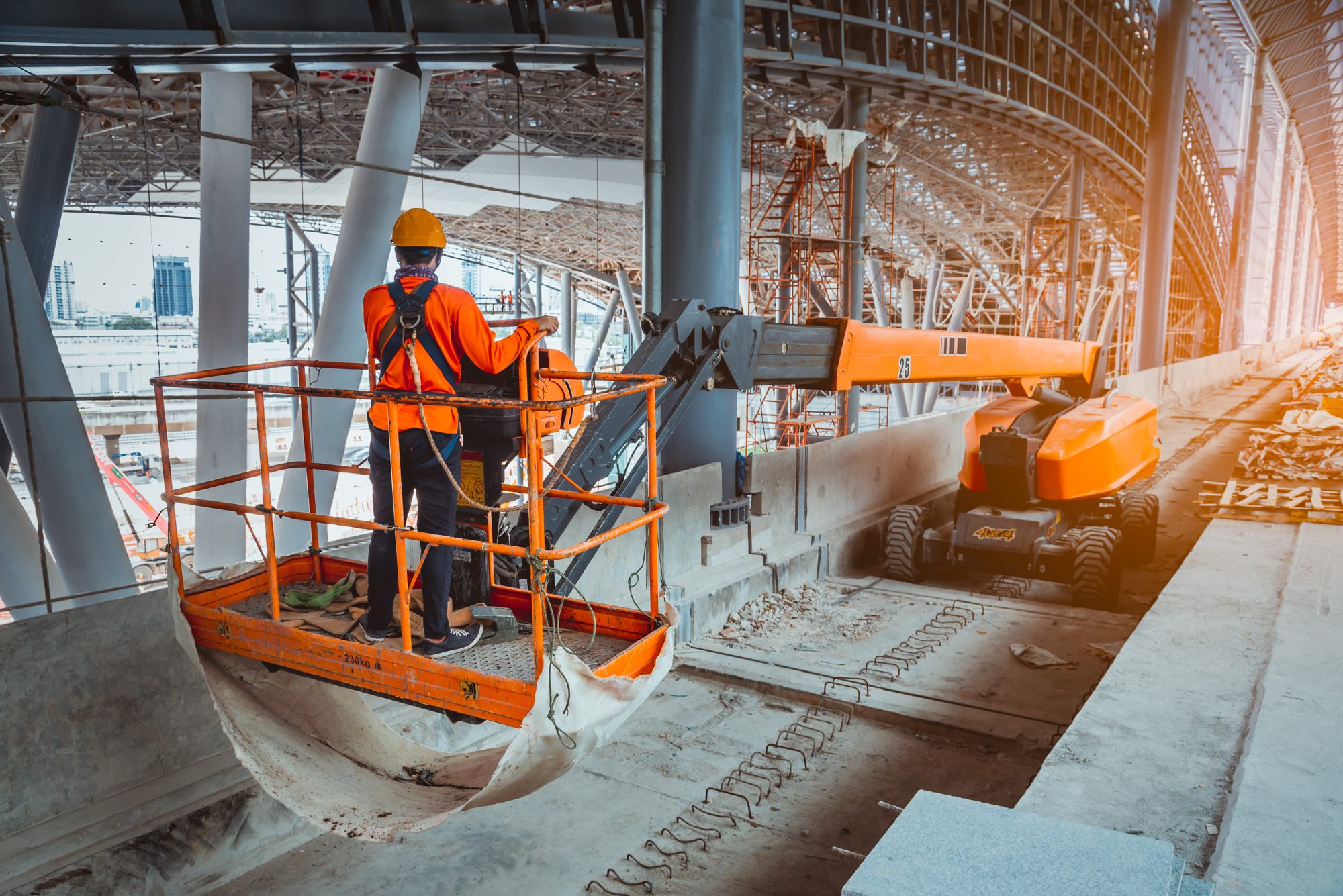 A worker in a safety harness on the platform of an orange boom lift at a construction site