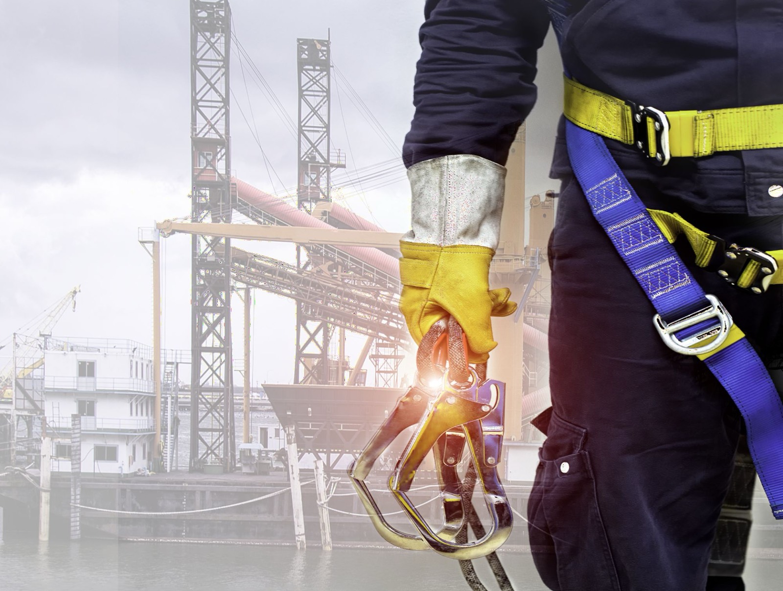 A worker in a safety harness holding two carabiners at an industrial site